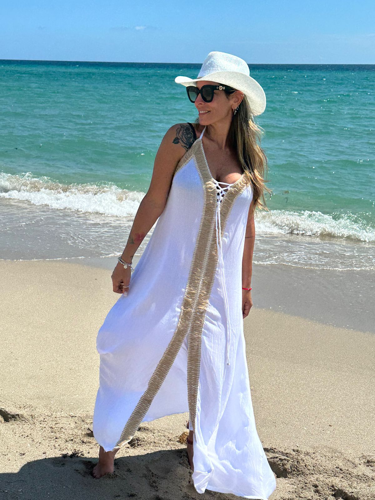 Woman walkingon the beach wearing a long white summer dress, sun hat and sunglasess, with the ocean in the background