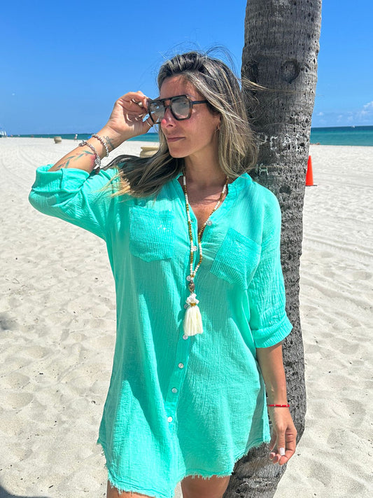 Model wearing short green cotton shirt dress with buttons, long sleeves and sequin details, beachwear style