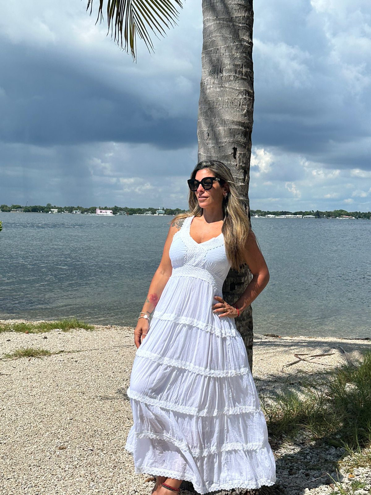 Model wearing the Ruby Dress, a long white cotton maxi dress, standing by the beach with palm trees