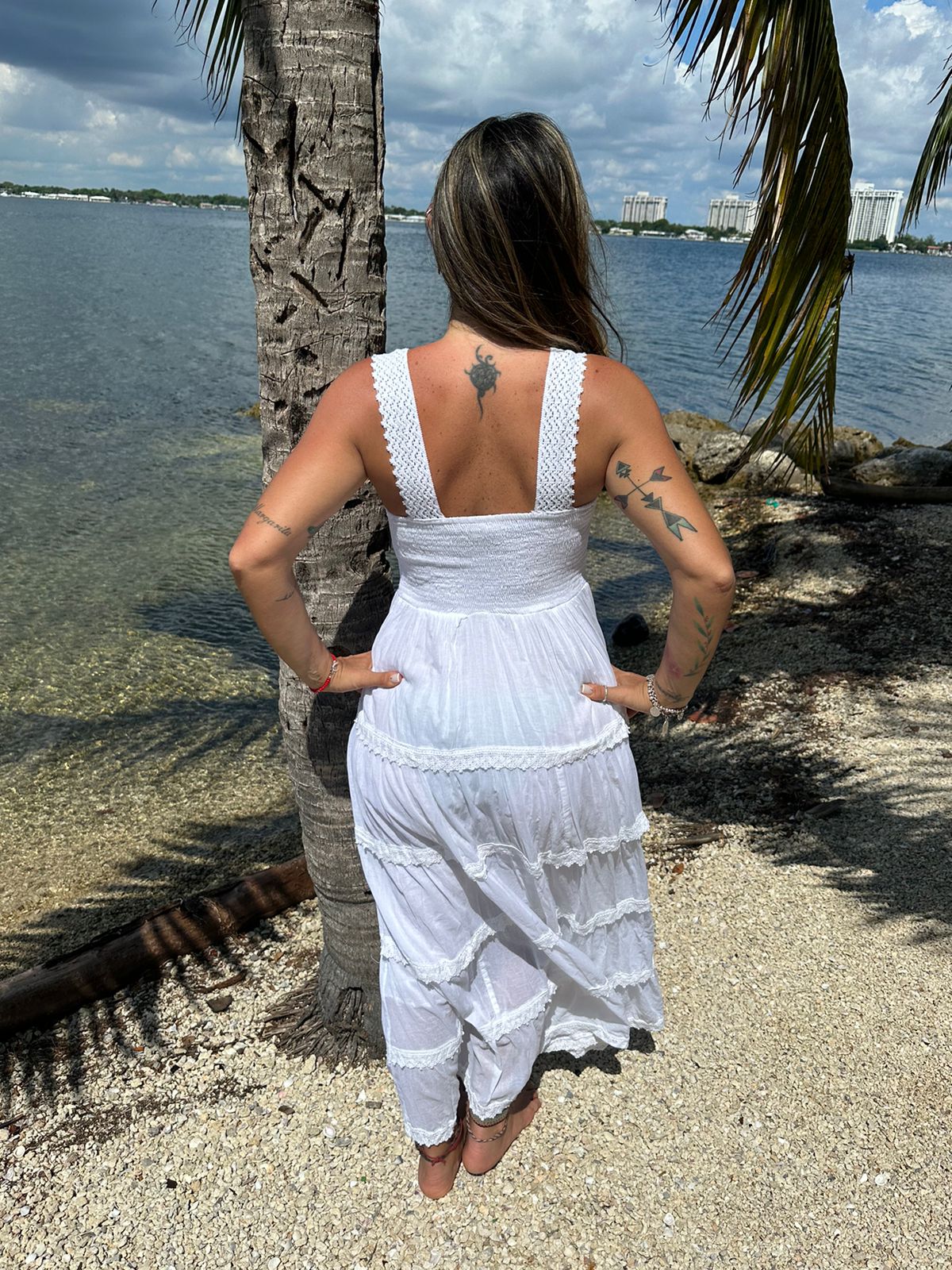 Back view of model wearing the Ruby Dress, a long white cotton maxi dress, standing by the beach with palm trees